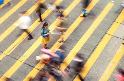 A woman pausing to focus on a message on her phone while on a busy crosswalk, with motion blur as people move around her. © Getty Images