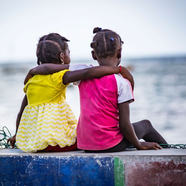 Two girls sit arm in arm on a wall facing the sea, Haiti. ©UNICEF/UNI274175/Bradley