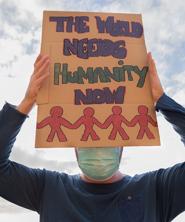 A masked person holding a sign in front of their face that reads: The world needs humanity now