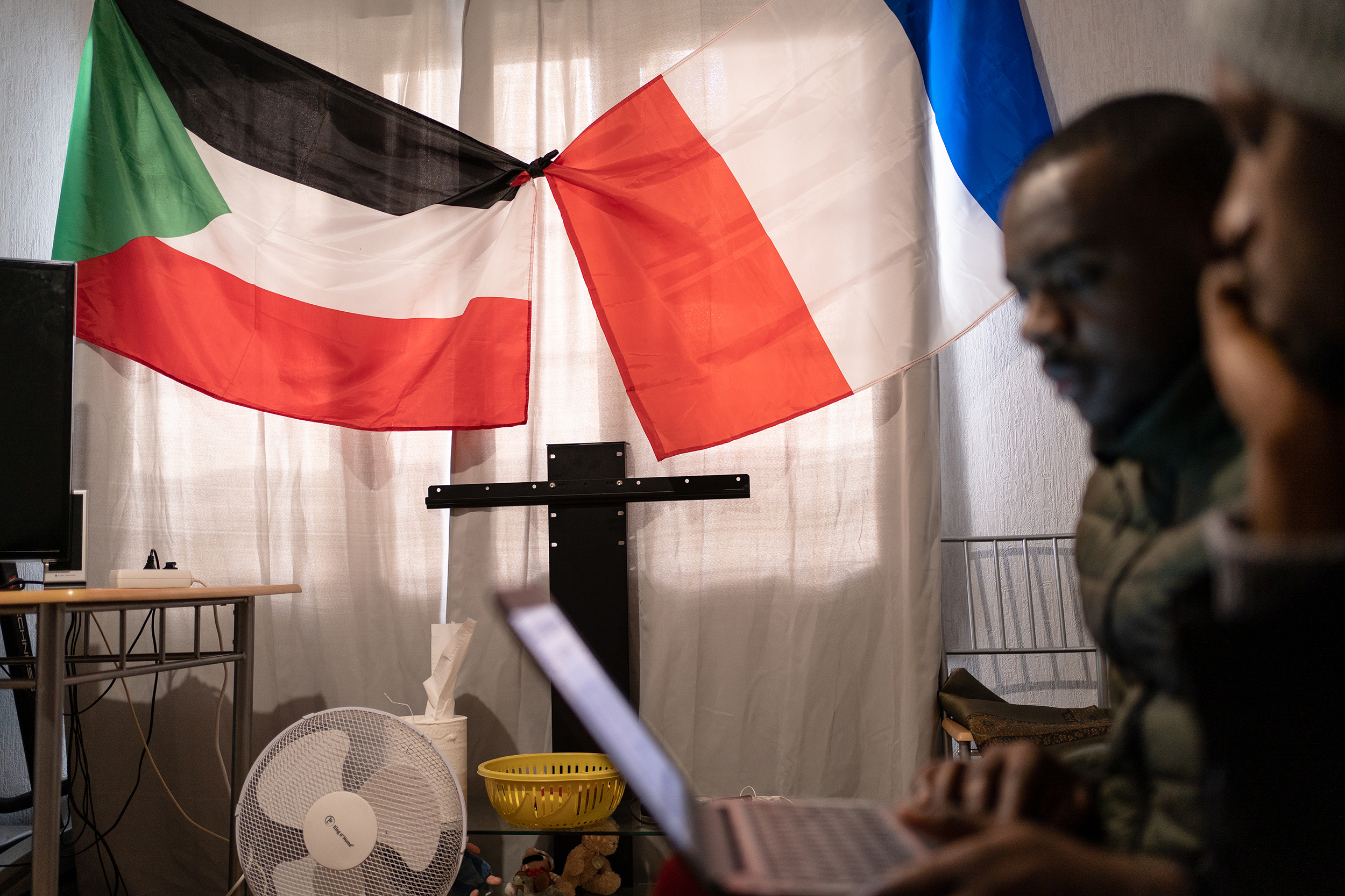 A man and a woman sitting in front of a laptop, with the flags of Sudan and France in the background