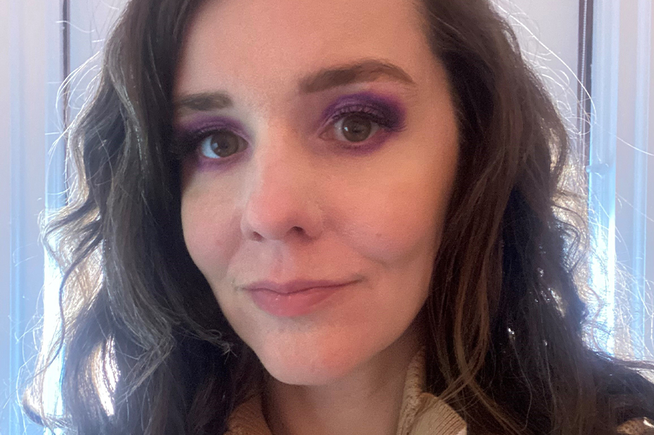 Portrait of Caroline Mazel-Carlton. She has long, wavy dark hair and wears bright purple eyeshadow around her brown eyes. © Caroline Mazel-Carlton