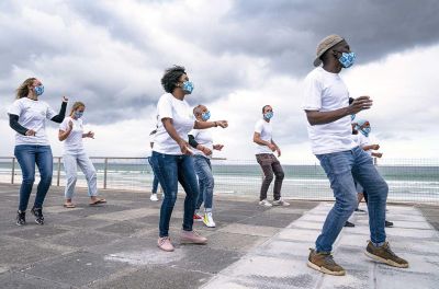 A group of young people of mixed race and gender and wearing jeans, white t-shirts and protective face masks perform a dance next to a beach.