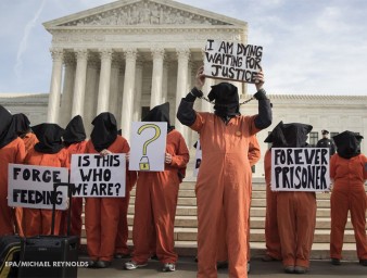 Protesters stand before a building, wearing orange jumpsuits and holding placards.