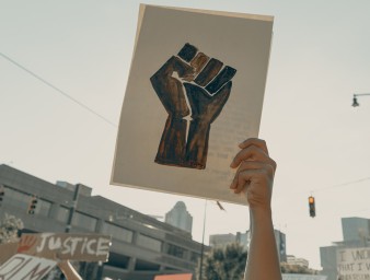 A woman in a crowd holds a demonstration sign depicting a balled fist, Charlotte, NC, USA, 3 June 2020 © Clay banks/UNSPLASH