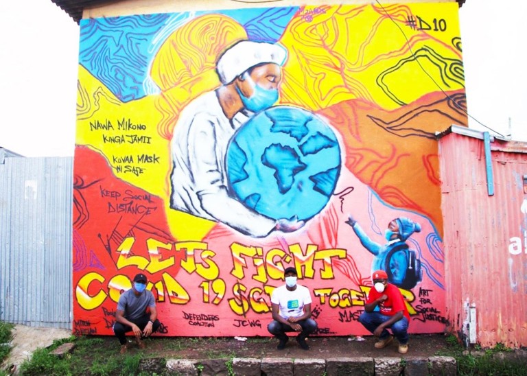 Three men in protective facemasks squat in front of a large wall mural depicting a health worker holding the globe and text that reads ‘Let’s fight COVID 19 together’.